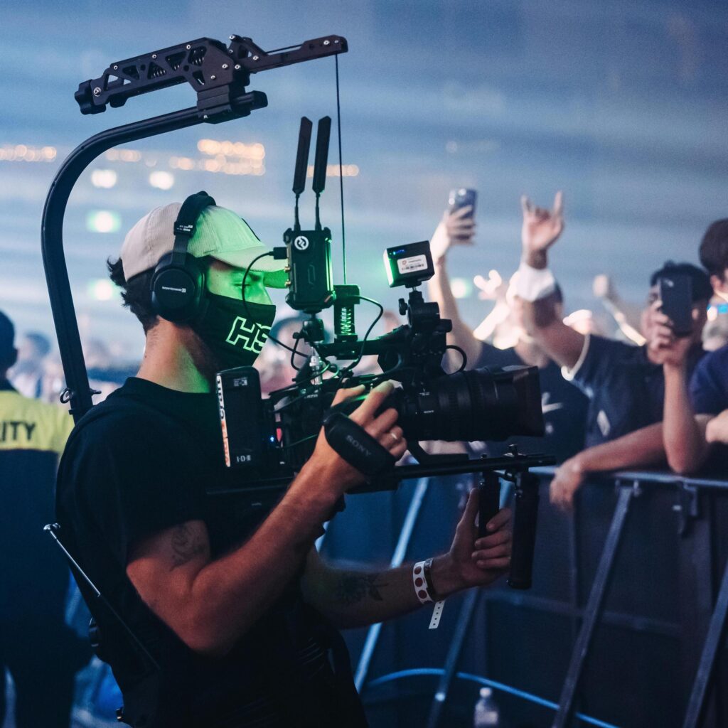 Cameraman with professional broadcasting equipment filming an event with a crowd in the background for broadcast services.